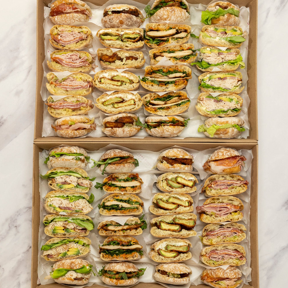 Two catering boxes filled with rows of various deli sandwiches, including meat, chicken, and avocado.