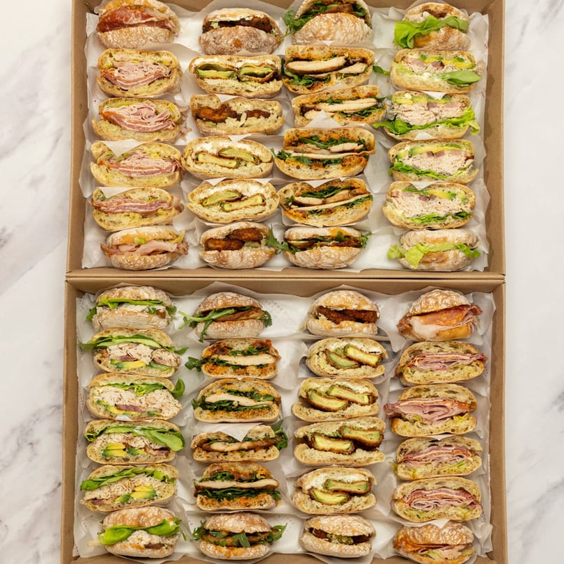 Two catering boxes filled with rows of various deli sandwiches, including meat, chicken, and avocado.
