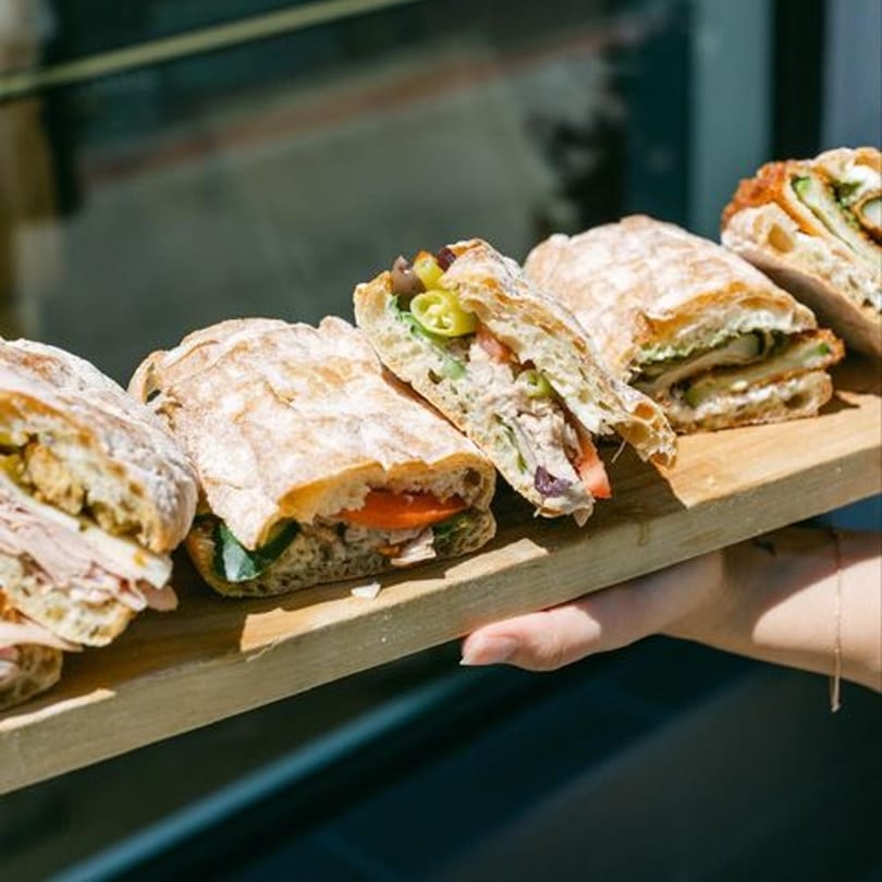 A hand holds a wooden board with five deli sandwiches on ciabatta bread.