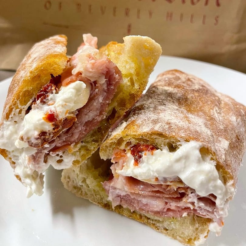 A deli sandwich from The Cheese Store of Beverly Hills, cut in half, revealing meat, cheese, and sun-dried tomatoes.