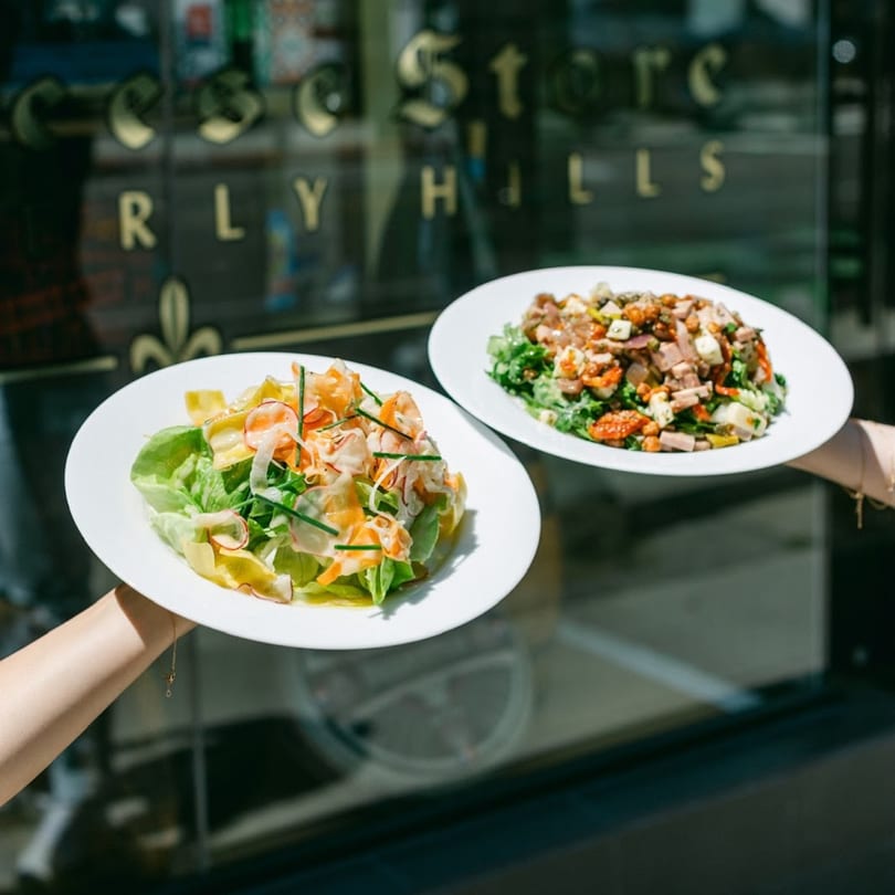 Two hands holding white plates with fresh salads in front of The Cheese Store of Beverly Hills.