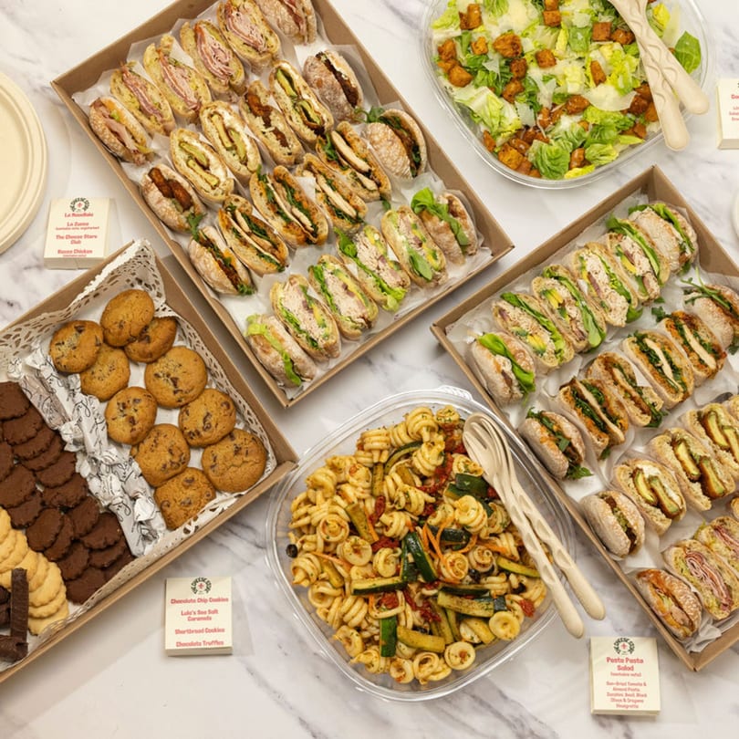 Deli catering spread with mini sandwiches, pasta salad, green salad, and dessert cookies.