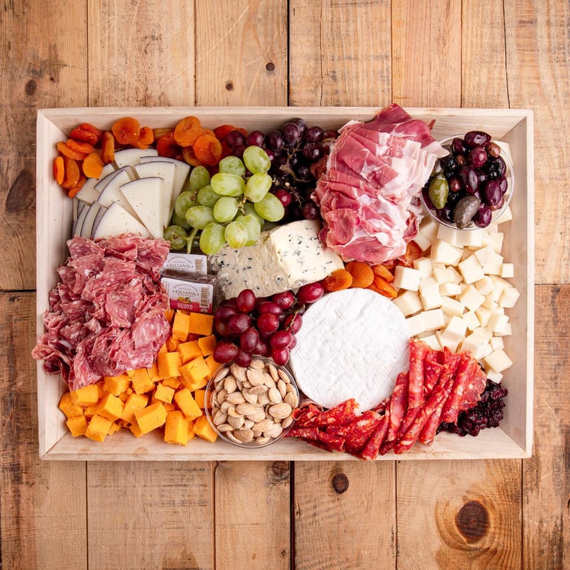 A wooden tray filled with various cheeses, meats, grapes, dried apricots, olives, and nuts.