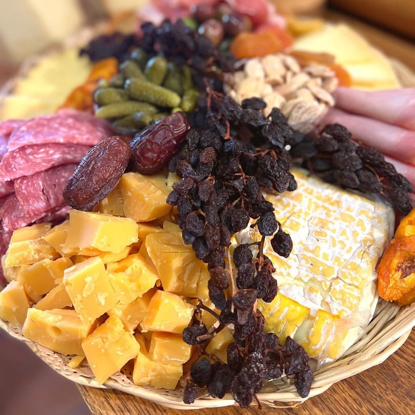 A deli platter with cheeses, meats, dried fruits, nuts, and pickles in a woven basket.